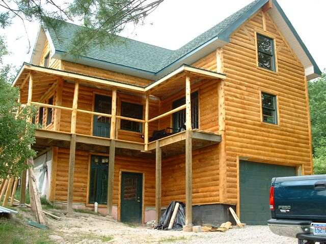 two story cottage finished in white cedar log siding, with a covered upper porch and a garage door below