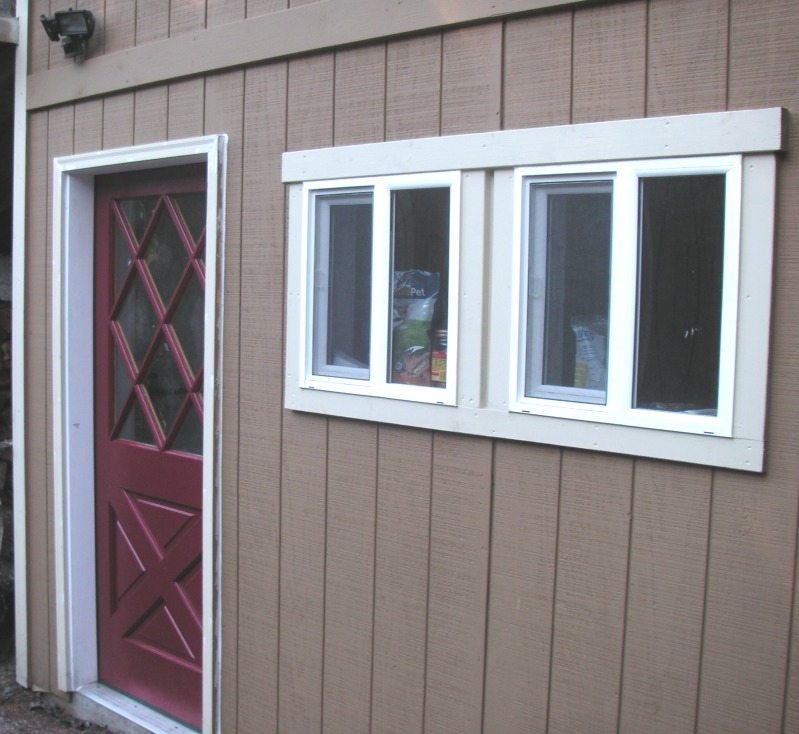two windows in the siding trimmed with plain flat boards and a wide head board, beside the red door
