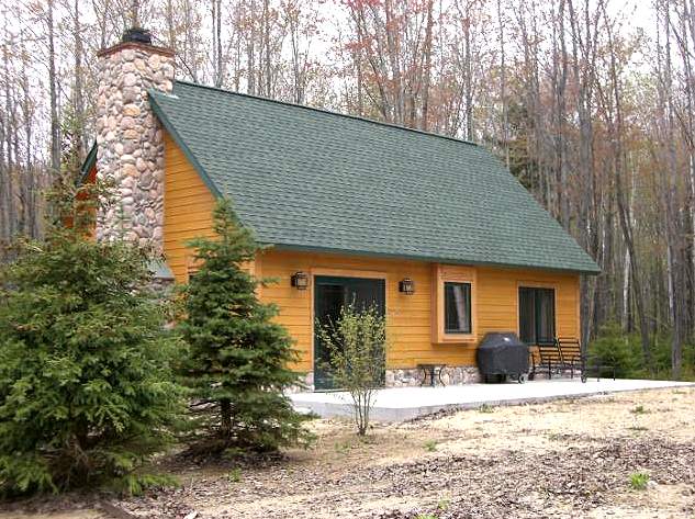 finished cabin with a green shingled roof, cedar colored siding and a river rock chimney, seen from the patio side