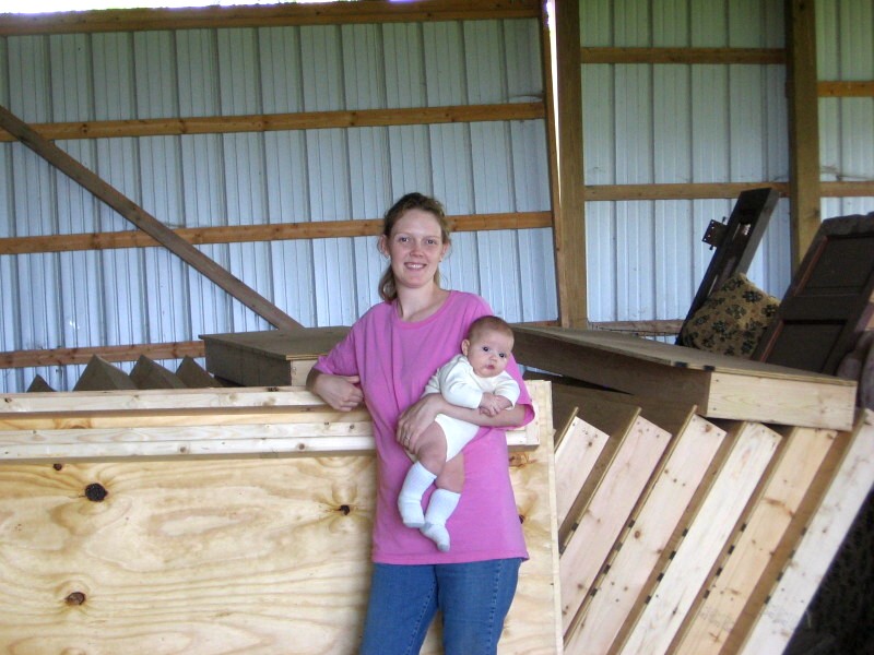 Cody holding her baby beside the stack of wall panels she framed in the barn