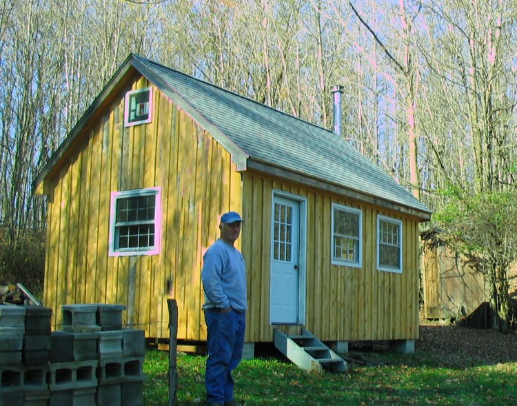 The same cabin two seasons on, now clad in pale board and batten siding with white trimmed windows and a white door, a man in a blue sweatshirt standing beside it and a stack of concrete blocks in the foreground.