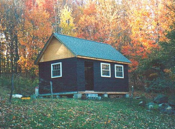 A small dark sided cabin with a green shingled gable roof and white trimmed windows, standing on block piers in a clearing surrounded by autumn maples.