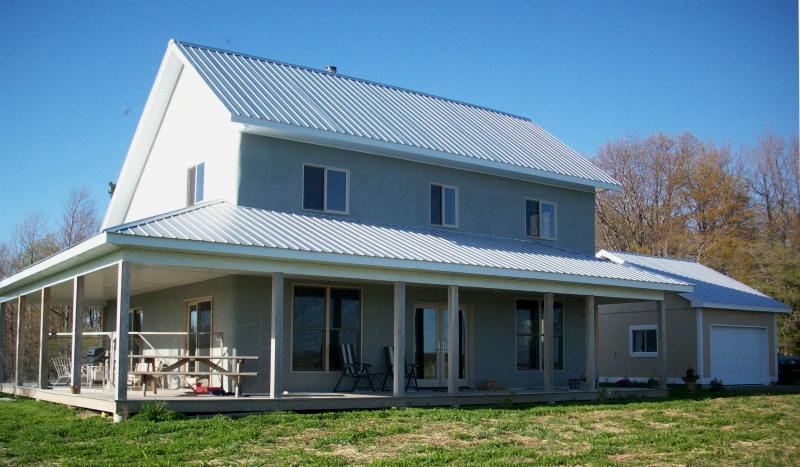 two story straw bale house with a metal roof and a covered porch around the front and side