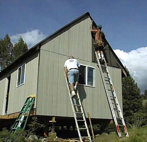 two builders on ladders nailing the T1-11 siding to the gable end of the cabin