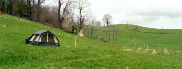 a tent pitched on a green hillside with stakes and batter boards laid out for the house