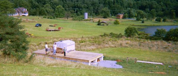 the framing package stacked on the newly decked first floor above the block foundation, with a pond and pasture beyond