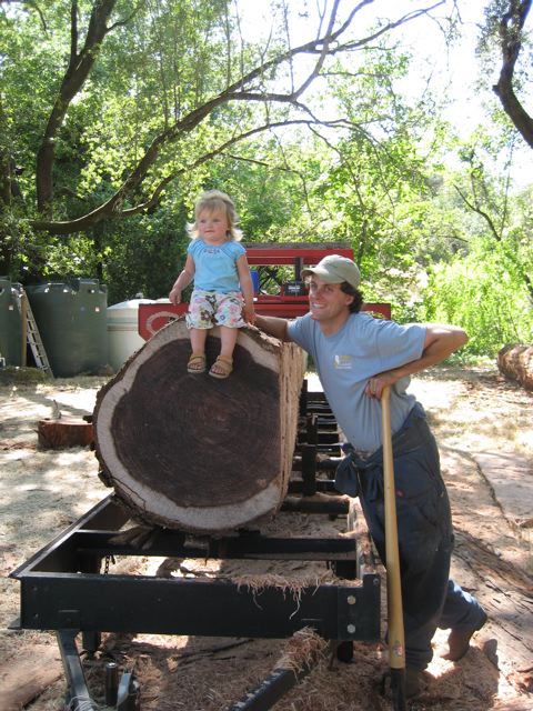 a young child sitting on a big redwood log on the portable sawmill, one of the builders alongside