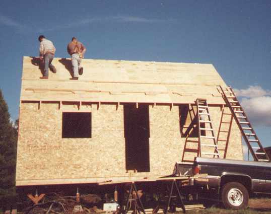 two builders sheathing the steep 12 in 12 roof with ladders leaning on the gable end